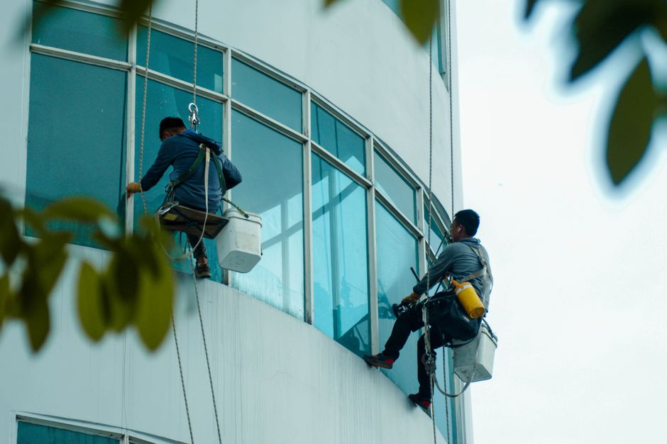 Two professional window cleaners from Snaresbrook Carpet Cleaners are performing surface cleaning on the exterior glass windows of a modern, multi-story building. They are suspended using safety harne
